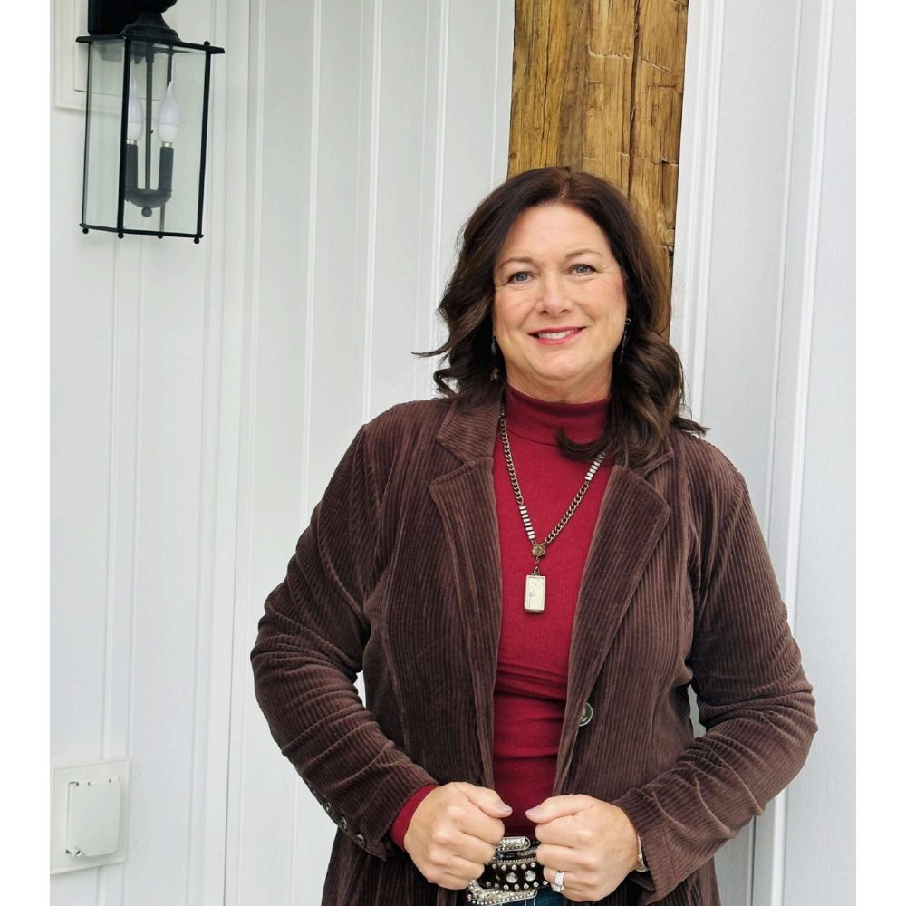 A woman in a brown jacket posing outdoors near a white wall.